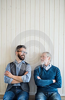 A portrait of adult hipster son and senior father sitting on floor indoors at home.