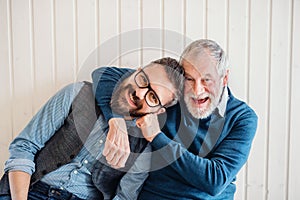 A portrait of adult hipster son and senior father sitting on floor indoors at home.