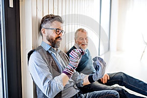 Portrait of adult hipster son and senior father sitting on floor indoors at home, having fun.