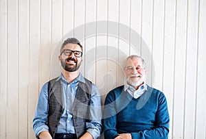 A portrait of adult hipster son and senior father sitting on floor indoors at home.