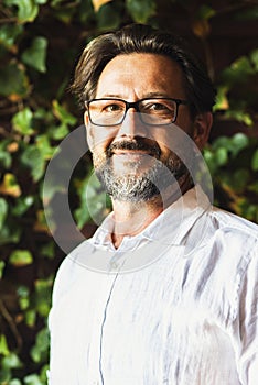 Portrait of adult mature handsome bearded man with white shirt smiling at the camera and wear eyewear. Foliage leaf green