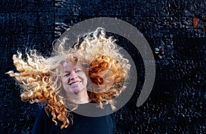 Portrait of adult mature happy laughing woman in front of burnt black, wooden wall shaking her long curly red hair