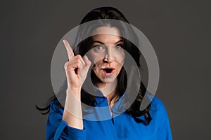 Portrait of adult thinking woman having idea moment pointing finger up on grey studio background. Smiling happy lady