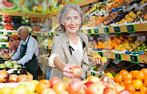 Portrait of adult woman choosing sweet apples and other fruit on counter of market