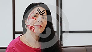 Portrait of adult woman with kinesio tapes on cheek and forehead, she gently touches her face with her fingers in medical clinic