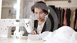 Portrait of an adult woman seamstress works at a sewing machine, working on a white hand made dress. Fashion clothes on