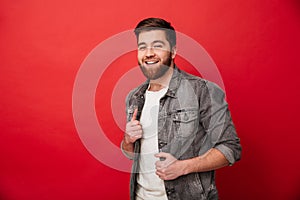 Portrait of affable kind man 30s in jeans jacket posing on camera with amazing smile, isolated over red background