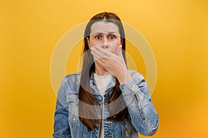 Portrait of afraid shocked young caucasian woman 25s wearing denim jacket covering mouth with palm, keep secret, does not tell