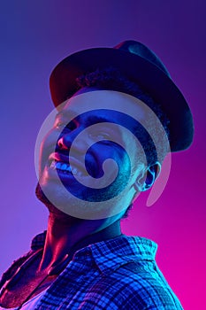 Portrait of African-American artist, Jazzman in hat smiling looking away in neon light against gradient blue-pink