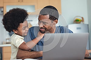 Portrait of African american cute little boy hugging of handsome dad working laptop computer while sitting at kitchen table at