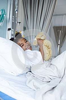 Portrait of african american girl patient lying on bed with teddy bear in patient room at hospital