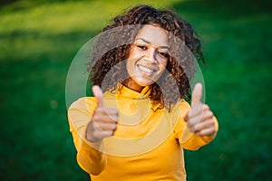 Portrait of african american girl showing thumbs up like gesture in green park. Young woman with curly hairstyle in
