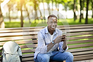 Portrait of African American guy using mobile phone on bench at summer park
