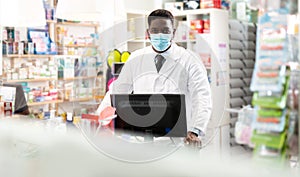 Portrait of an african american male pharmacist in a protective mask, standing behind the counter of a pharmacy
