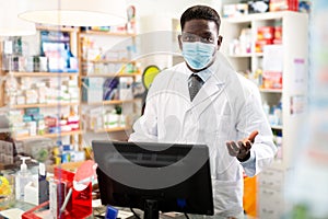 Portrait of an african american male pharmacist in a protective mask, standing behind the counter of a pharmacy