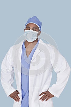 Portrait of an African American male surgeon with hands on hips over light blue background