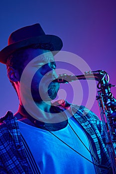 Portrait of African-American man, focused musician playing saxophone in hat against blue and pink background in neon