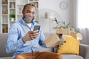 Portrait of an African-American man sitting on the sofa at home, holding an open parcel box in his hand, using a mobile