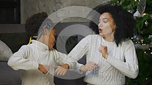 Portrait of African American mom and her little daughter funny dancing and smiling. Woman and girl sitting near sofa and