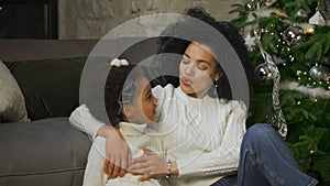 Portrait of African American mom and her little daughter hugging and talking cute. Woman and girl sitting near sofa and