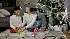 Portrait of African American mom and her little daughter tying a bow on a gift box. Woman and girl sitting near sofa and