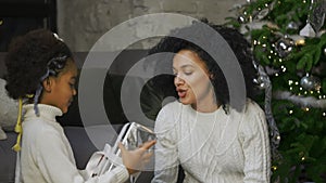 Portrait of African American mom surprises her little daughter with a gift. Woman and girl sitting near sofa and