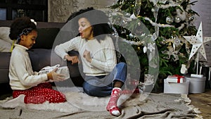 Portrait of African American mom surprises her little daughter with a gift. Woman and girl sitting near sofa and