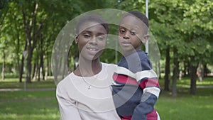 Portrait of African American woman holding her son in her arms in the green park close up. Cute child spending time