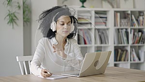 Portrait of African American woman talking on video conference call using laptop and headphones taking notes on notepad