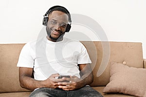 Portrait of an african american young guy enjoying listening to good music at home, sitting on the sofa, looking at