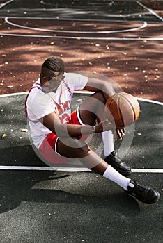 Portrait of an African American young man holding a basketball and sitting on a basketball court. Take a break during