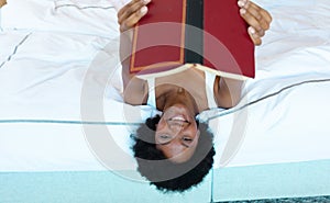 Portrait of african american young woman smiling while holding a book lying on the bed at home