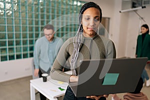Portrait of African female entrepreneur standing in modern office holding laptop computer, looking to screen. Diverse