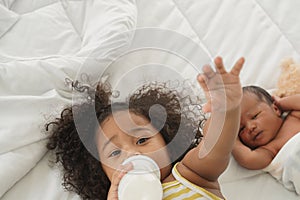 Portrait of African little girl is lying in bed and sucking a milk bottle with her newborn baby boy and looking at camera