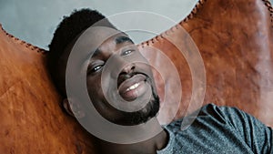 Portrait of African man sitting in chair, smiling and looking straight at camera. Male looks dreamy, thoughtful and calm