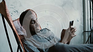 Portrait of African man sitting in chair, using a Smartphone. Handsome male smiles and looks at photos in his phone.