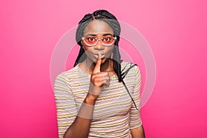 Portrait of african woman holding index finger on lips and asking to keep silence isolated over pink background