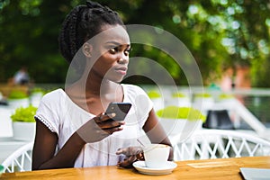 Portrait of african woman using mobile phone at an outdoor cafe with laptop and cup of coffee on table