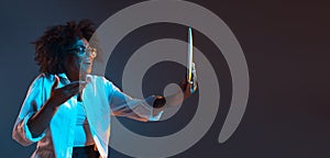 Portrait of African young girl in white shirt using tablet, gadget isolated on dark blue studio background. Concept of