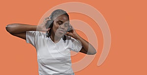 Portrait of African young woman listening to music on headphones, eyes closed and holding headphones with hands against orange