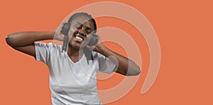 Portrait of african young woman listening to music on headphones with toothy smile, holding headphones with hands against orange