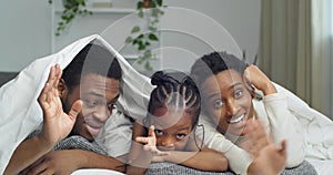 Portrait of afro american ethnic black mixed race family lying covered with white blanket waving at camera hello smiling