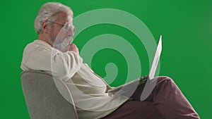Portrait of aged bearded man on chroma key green screen. Senior man in casual clothing sitting on chair and working on