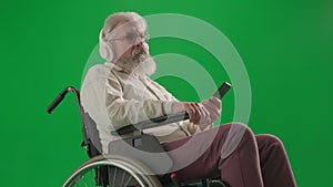 Portrait of aged disabled man on chroma key green screen. Close up senior man in wheelchair in headphones holding