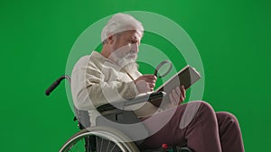 Portrait of aged disabled man on chroma key green screen. Close up senior man in wheelchair reading book with magnifying