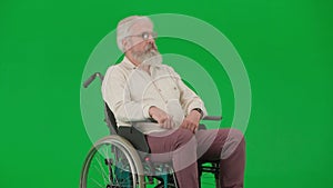 Portrait of aged disabled man on chroma key green screen. Elderly man sits in wheelchair, looks away, then smiles at the