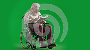 Portrait of aged disabled man on chroma key green screen. Full shot senior man in wheelchair reading book with