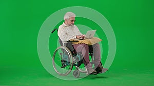 Portrait of aged disabled man on chroma key green screen. Senior man sitting in wheelchair with laptop on the pillow