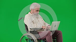 Portrait of aged disabled man on chroma key green screen. Senior man sitting in wheelchair and working on laptop, typing