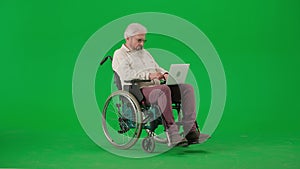 Portrait of aged disabled man on chroma key green screen. Senior man sitting in wheelchair and working on laptop, typing